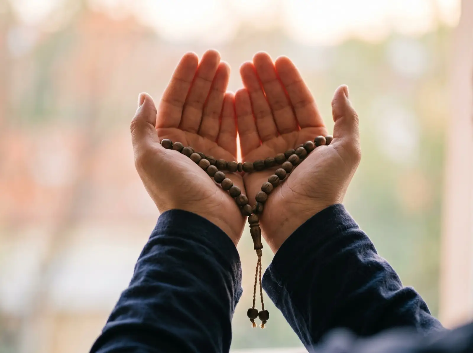 A pair of open hands raised in supplication with wooden prayer beads resting across the fingers, back-lit by soft warm light.