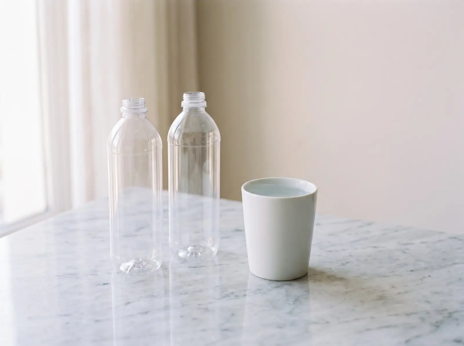 Two clear water bottles and a white cup of Zamzam water standing on pale polished marble in soft daylight.
