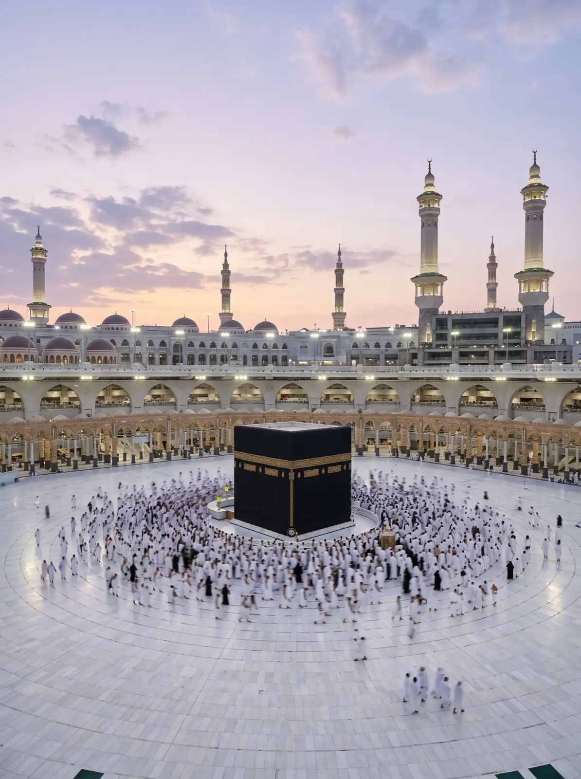 Pilgrims in white ihram making tawaf around the Kaaba in the courtyard of Masjid al-Haram at dawn, under a pale lavender sky.